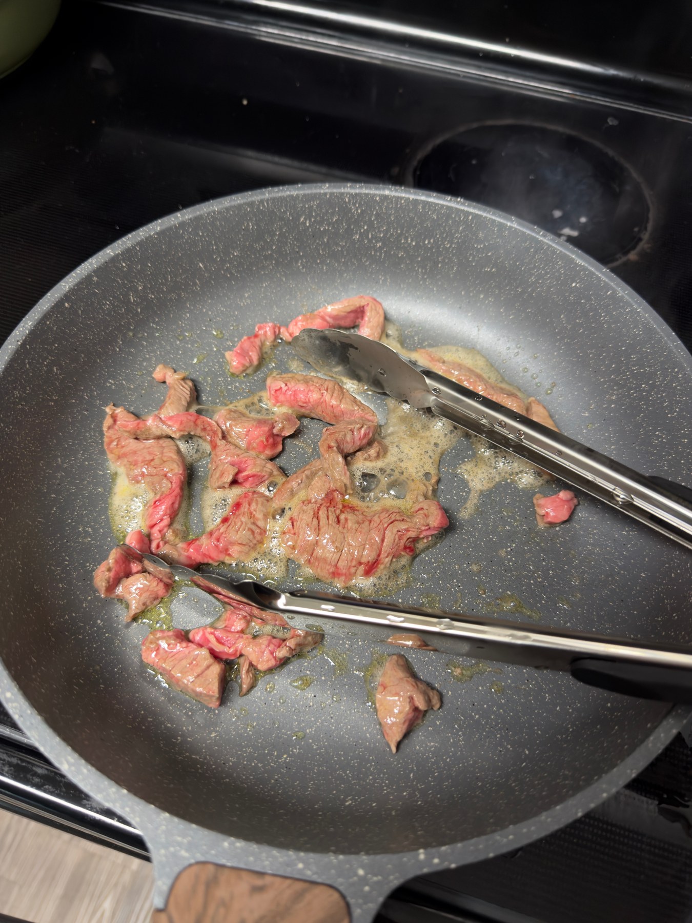 Sliced meat and metal tongs in a gray speckled frying pan with a wood-look handle, on a black electric stovetop.